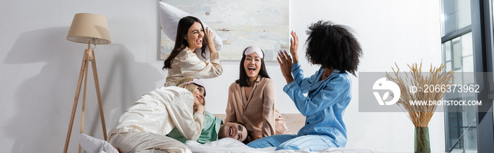 group of cheerful interracial friends in pajamas having pillow fight during slumber party, banner