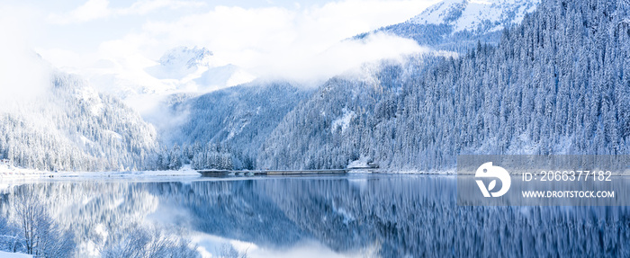 Panoramic view of beautiful white winter wonderland forest scenery in the Alps with snowy mountain summits reflecting in crystal clear mountain lake on a cold sunny day with blue sky and clouds