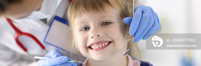 Dentist doctor examines teeth of little smiling girl in clinic. Painless dental treatment for children concept.