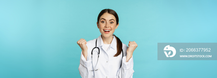 Happy and excited young female doctor, physician celebrating, achieve goal and smiling pleased, winning, standing in white coat against blue background