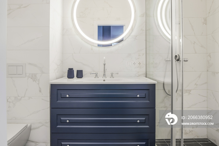 Modern bathroom interior with a dark blue wooden shelf, white sink and big round mirror with LED lig