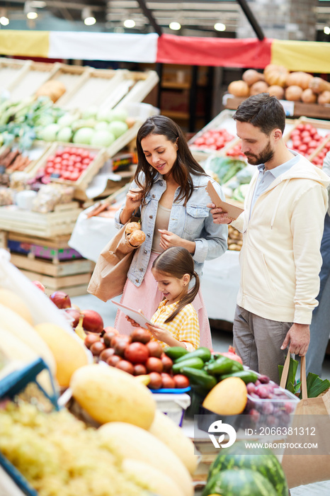 Positive young family standing at food shelves and using online shopping app for purchasing on table