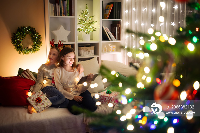 Two cute young sisters using a tablet pc at home in warm and cozy living room at Christmas. Family h