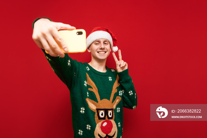 Happy young man in christmas clothes taking selfies on smartphone, looking at camera phone and showi