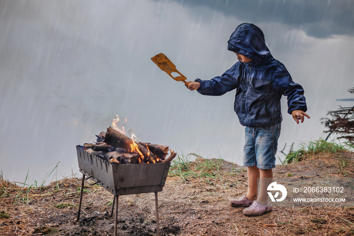 Boy in wet jacket barbequing and warming arms near open fire in rain, family autumn weekend