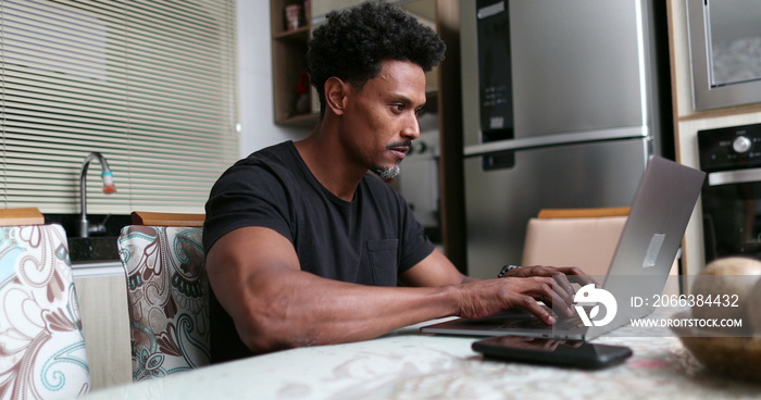 African man typing on computer laptop at home kitchen