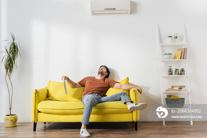 man holding newspaper while lying on yellow couch and suffering from heat in living room