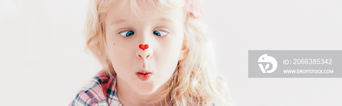 Cross-eyed kid looking at red heart sticker on her nose. Funny hilarious white Caucasian cute adorab