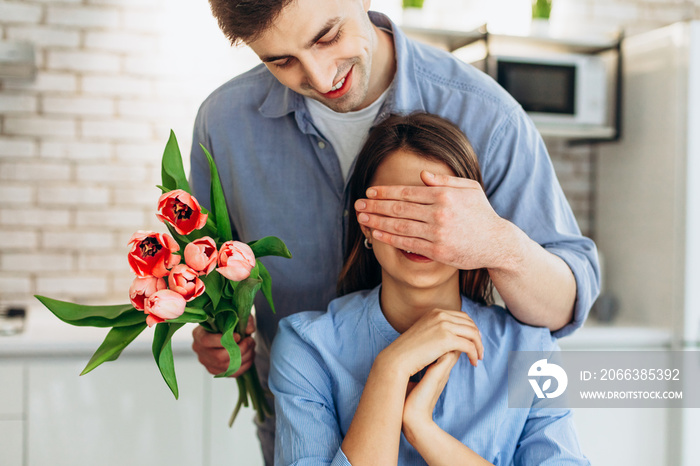 Surprise! Beautiful romantic couple at home. Young man is presenting flowers to his beloved. Feel of