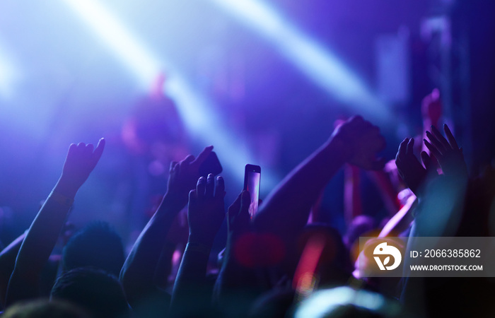 Audience with hands raised at a music festival and lights streaming down from above the stage. Crowd