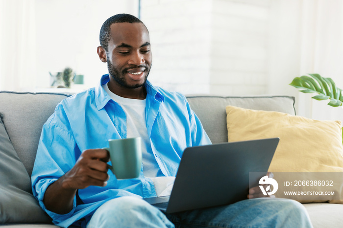 Portrait of smiling black man using laptop, holding cup and drinking morning coffee, sitting on sofa