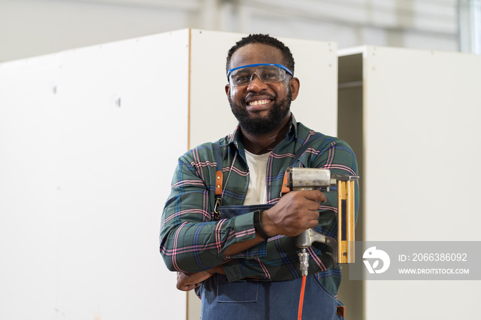 African American male carpenter holding pneumatic nail gun at wood workshop. Portrait of joiner male