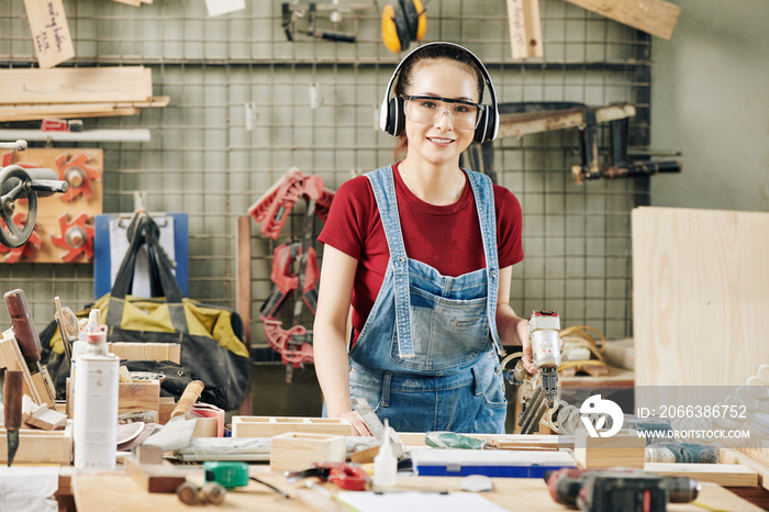 Portrait of pretty young female carpenter in earmuffs and goggles working with pneumatic stapler whe