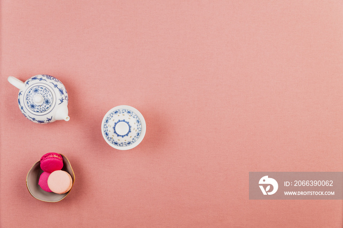 Chinaware teapot and bowl over a pink tablecloth background with pink french macarons in a bowl with large copyspace.