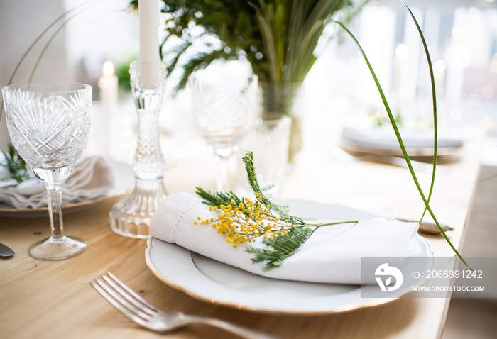 Beautiful springtime table setting with green leaves and mimosa branches, bright white table dinner decoration