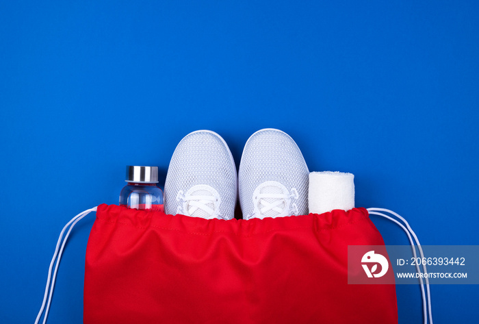 White sport shoes sneakers, glass water bottle and towel in red sport bag on cyan blue background.