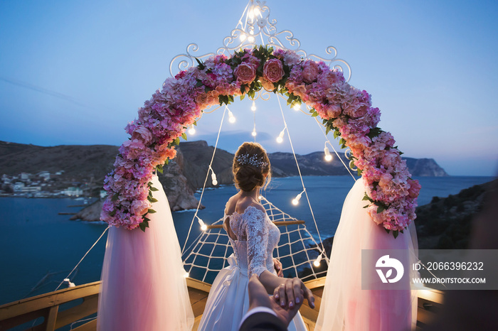 The bride holds the groom’s hand against the background of a wedding arch with bright lights at sunset on the seashore. The wedding ceremony.