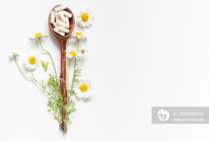 Herbal medicine concept. Wooden spoon with white medical capsules with fresh chamomile flowers on white background top view.