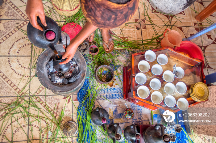 A woman with a visor and sandals seen from above while preparing a cup of coffee heated with charcoal in the typical Ethiopian coffee basket and with herbs scattered on a tiled floor and a tablecloth.