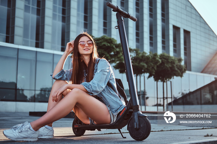 Beautiful young woman in sunglasses is sitting on her electro scooter near big glass building.