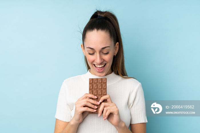 Young brunette woman over isolated blue background eating a chocolate tablet