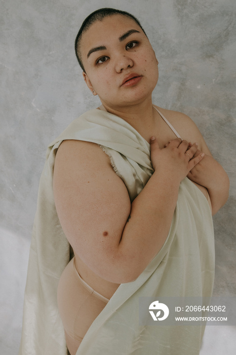 portrait of a plus size filipino woman with a shaved head and hands on her heart with satin fabric l