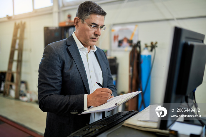 Mid adult manager using computer while taking notes in auto repair shop.