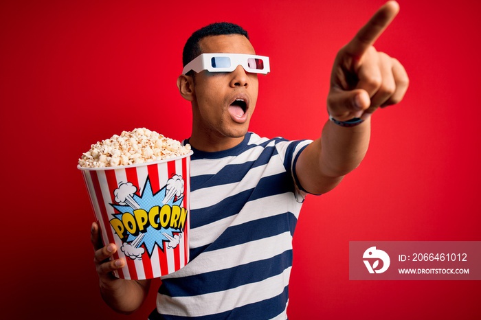 Young handsome african american man watching movie using 3d glasses eating popcorns Pointing with fi