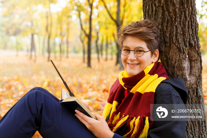Cute pretty boy in costume of Harry Potter and scarf plays as  a magician, reads book in autumn park
