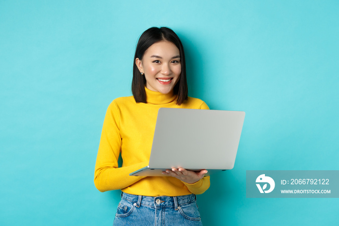 Happy asian woman in yellow pullover using laptop, shopping online or working, standing over blue ba