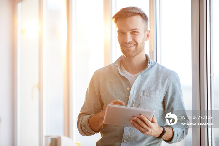 Waist up portrait of handsome young man holding digital tablet standing  by window in sunlight and l