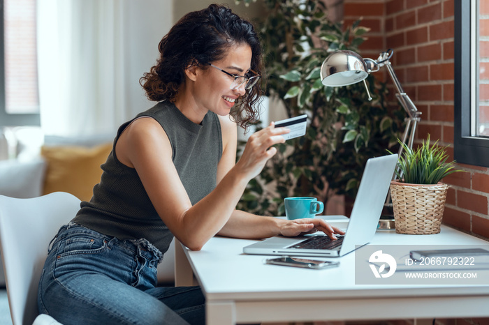 Cute woman holding white credit card for shopping online with computer while sitting in living room 