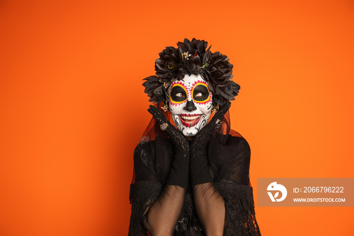 smiling woman in sugar skull makeup and black halloween costume holding hands near face isolated on 