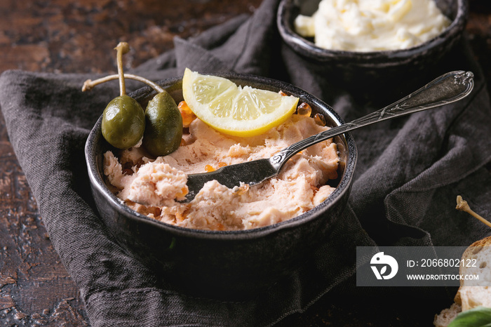 Black bowl of salmon pate with red caviar served with butter, sliced bread, capers, vintage knife and herbs on textile linen napkin over brown texture background. Close up