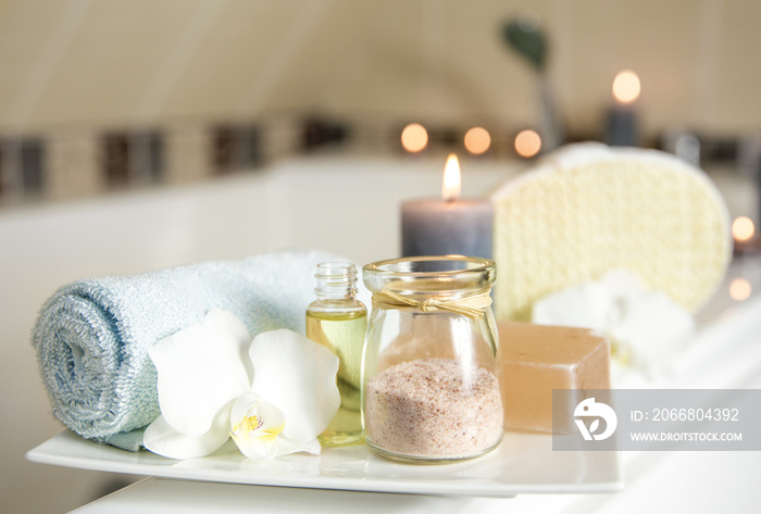 White ceramic tray with home spa supplies in home bathroom for relaxing rituals. Candlelight, salt soap bar, bath salt in jar, massage, bath oil in bottle, blue rolled towel, natural sponge.