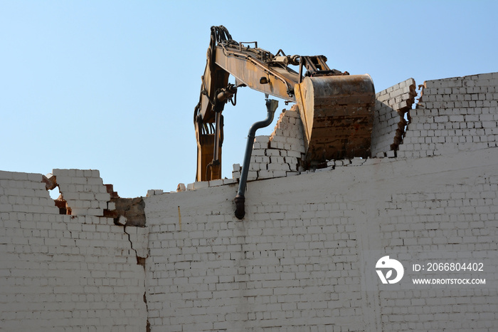 An excavator bucket is dismantling an old brick wall against the background of the sky