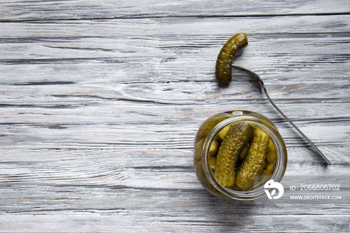 Open jar with pickled cucumbers and cucumber on a fork on a light wooden background top view.