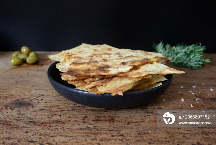 A photographic composition of a black dish on a wooden table with  pane carasau , a typical Italian bread. In the background, some green olives and aromatic herbs. Italian cuisine and gastronomy.