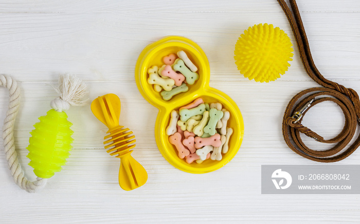 Yellow bowl with snacks, ball, leash, bone toy for dogs on white wooden table. Set of accessories for pets on wood. Close up