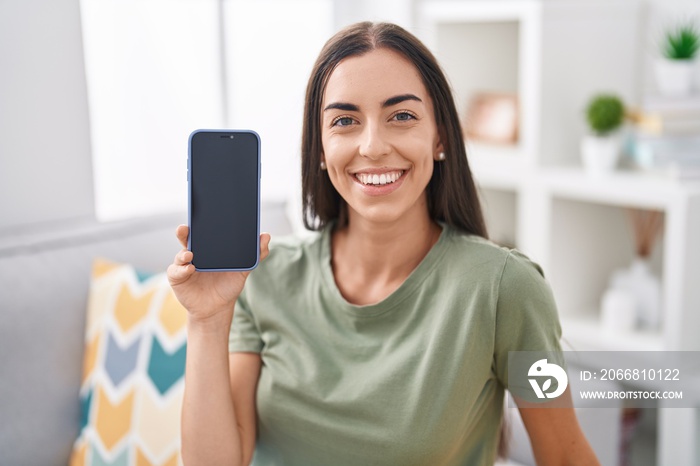 Young brunette woman holding smartphone showing blank screen looking positive and happy standing and smiling with a confident smile showing teeth