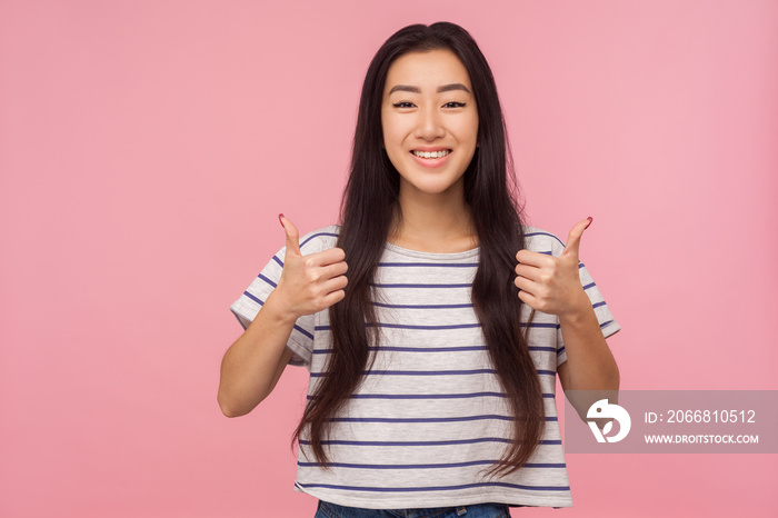 Well done, like it! Portrait of glad satisfied girl with long hair in striped t-shirt showing thumbs up, happy about successful project, excellent job. indoor studio shot isolated on pink background