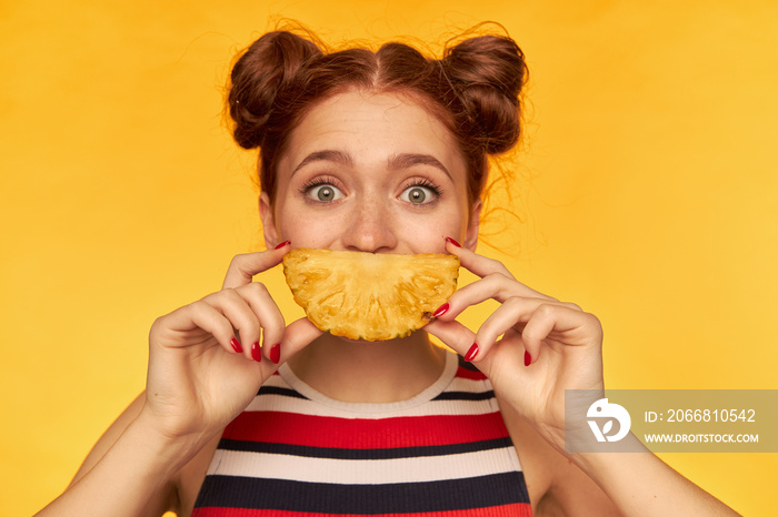 Teenage girl, happy looking red hair woman with two buns and big eyes. Wearing striped tank top and holding slice of a pineapple. Watching at the camera, isolated closeup over yellow background