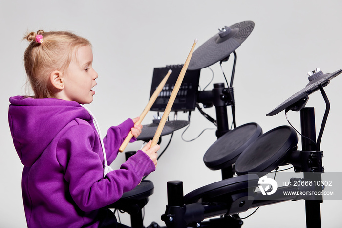 Happy caucasian kid girl drummer. Girl playing on elettronic drum kit or learns to play drums in music school. Emotional portrait. Mock up, studio on gray background. Free space for advertisement.