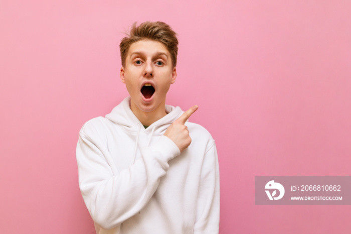 Portrait of surprised young man in casual clothes stands on pink background and shows thumbs aside, looks into camera with shocked face. Shocked guy points his finger at a blank spot.