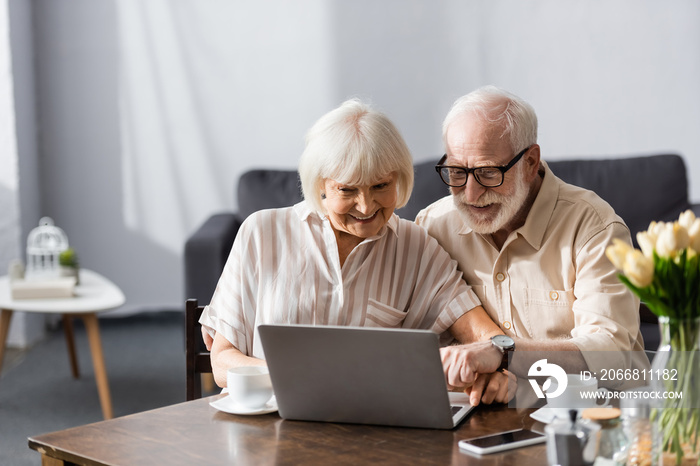 Selective focus of smiling elderly couple using laptop near coffee cups on table