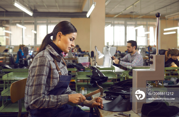 Portrait of concentrated skilled young caucasian woman shoemaker in apron and uniform working with leather and hammer at workplace side view. Shoe factory, footwear industry and manufactory