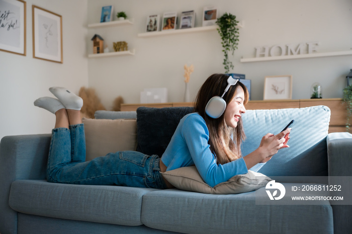Happy young asian woman relaxing at home. Female smile sitting on sofa and holding mobile smartphone. Girl using video call to friend. Listening to music
