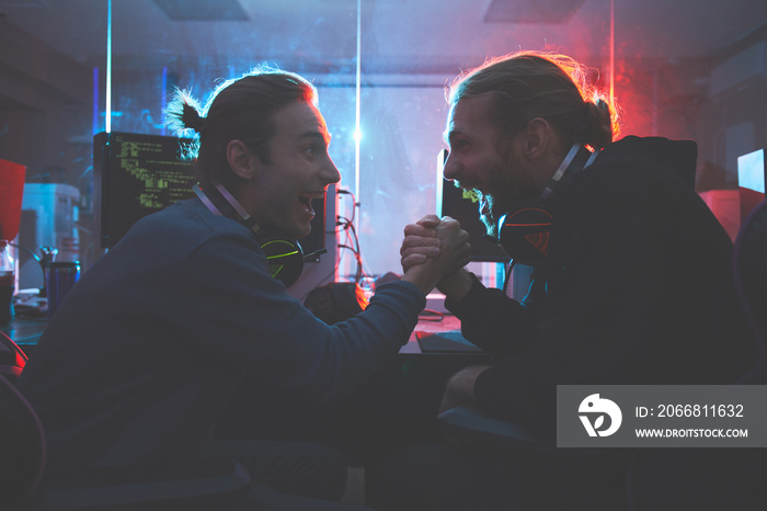 Cheerful young hipster software engineers with headsets on necks sitting in database center and shaking hands while being excited