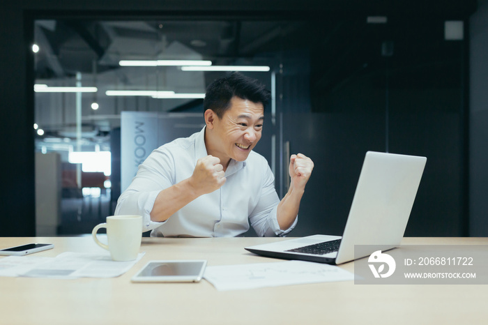 A young Asian man sits in the office at the table, during working hours he watches a football match, sports competitions,makes bets, cheers and rejoices on his laptop.