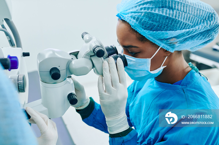 Young Black female surgeon in medical mask and rubber gloves looking through electronic miscroscope